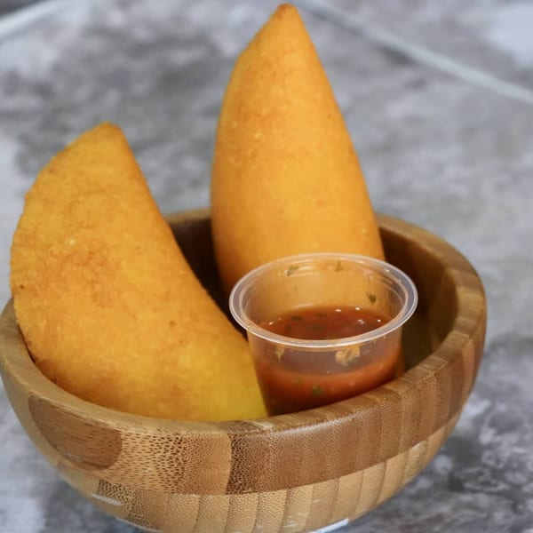 Two golden-fried Venezuelan empanadas and a small cup of red sauce in a wooden bowl.