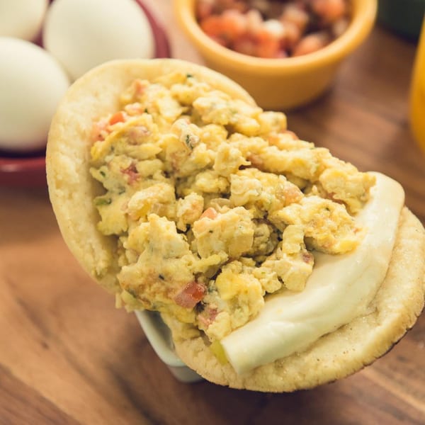 A Venezuelan arepa filled with scrambled eggs and white cheese, with eggs and salsa in the background.