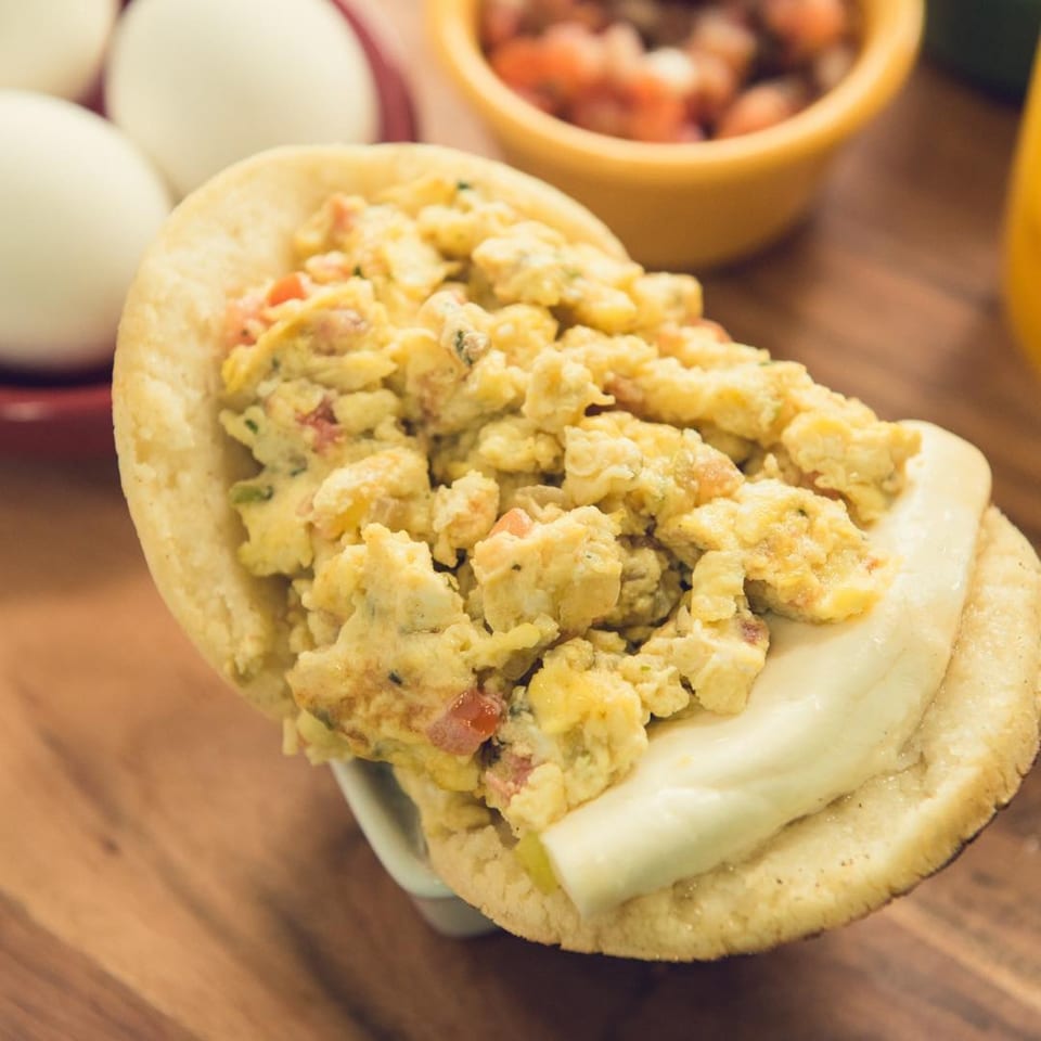 A Venezuelan arepa filled with scrambled eggs and white cheese, with eggs and salsa in the background.
