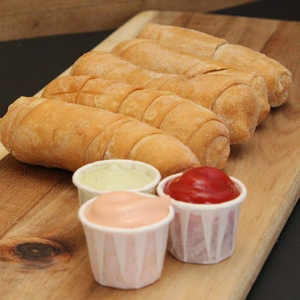 Golden-brown Venezuelan tequeños, a fried cheese pastry, arranged on a wooden board with three dipping sauces.